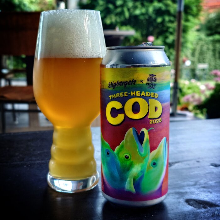 Ölburk med etikett "Three-Headed Cod 2025" bredvid ett glas fyllt med ljus öl, stående på ett bord utomhus.