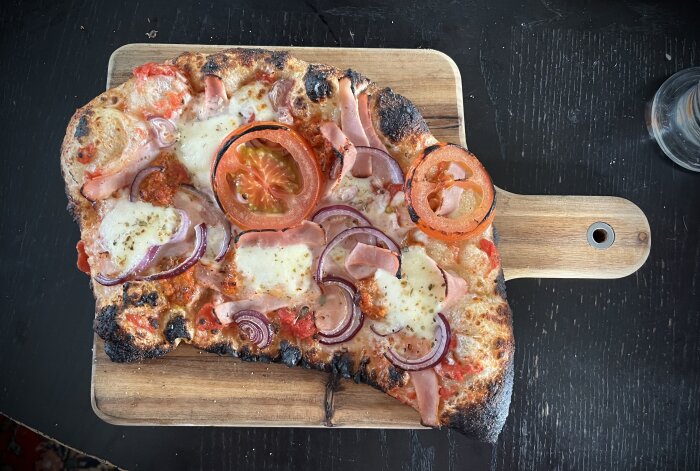 Hemlagad pizza med bränd kant, toppad med mozzarella, tomat, skinka och rödlök på en träskärbräda.