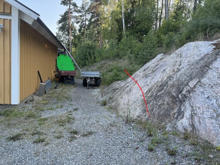 Gul byggnad bredvid grusväg, två släpvagnar och stor sten med röd markering visar planerat markarbete vid skogstomt.