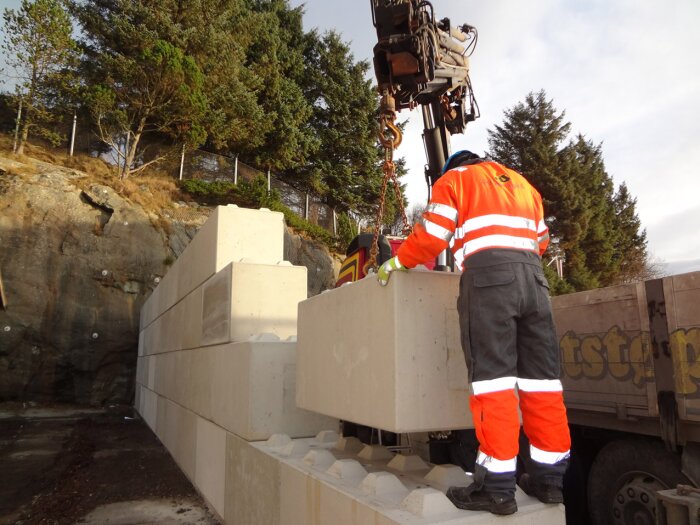 Arbetare i orange skyddskläder styr lyft av betongblock vid bergvägg. Grävmaskin används för att placera blocken i konstruktionen.