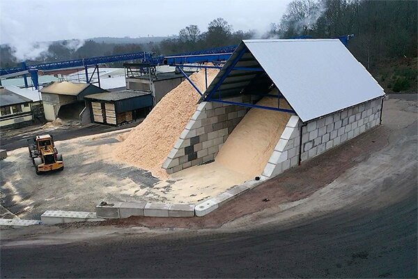 Industriell anläggning med stora högar sågspån under plåttak, omgiven av betongblock. En hjullastare syns vid sidan.