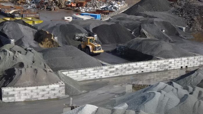Grävskopa i stenig miljö, omgiven av högar av grus och grusblock, med staplade betongblock i förgrunden och metallskrot i bakgrunden.