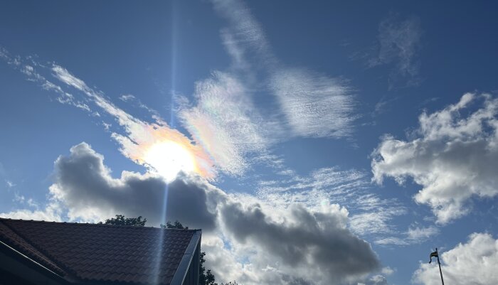 Blå himmel med moln och solens strålar skapar ett spektrum av färger. Del av hustak och flaggstång syns.