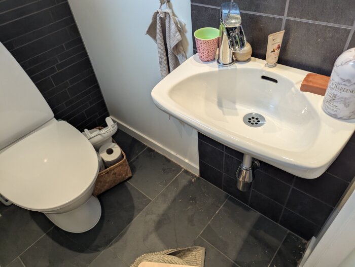 Badrum med toalett och handfat, svarta plattor på golvet, handduk hänger på väggen, korg med toalettpapper under handfatet.