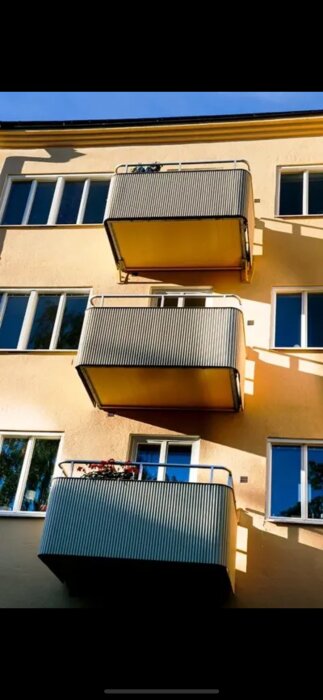 Flera grå balkonger i rad på en gul byggnadsfasad, med fönster och skuggor.