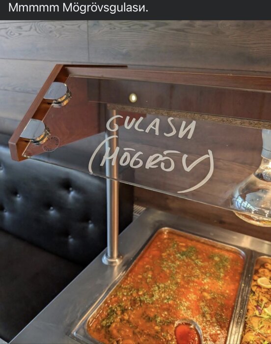 Buffé med soppa märkt "Gulasi (Mögröv)" i ett värmeskåp, del av serveringsområde med transparent skydd.