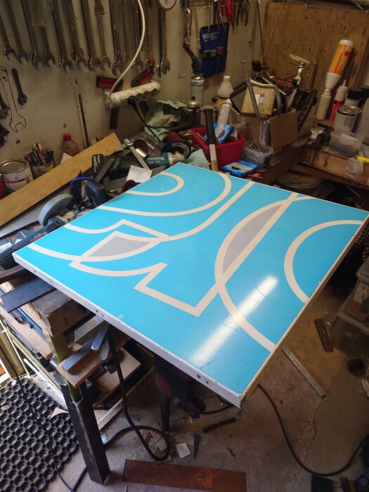 Blå skylt med vita grafiska element liggande på ett arbetsbord, omgiven av verktyg och material i ett verkstadsliknande utrymme.