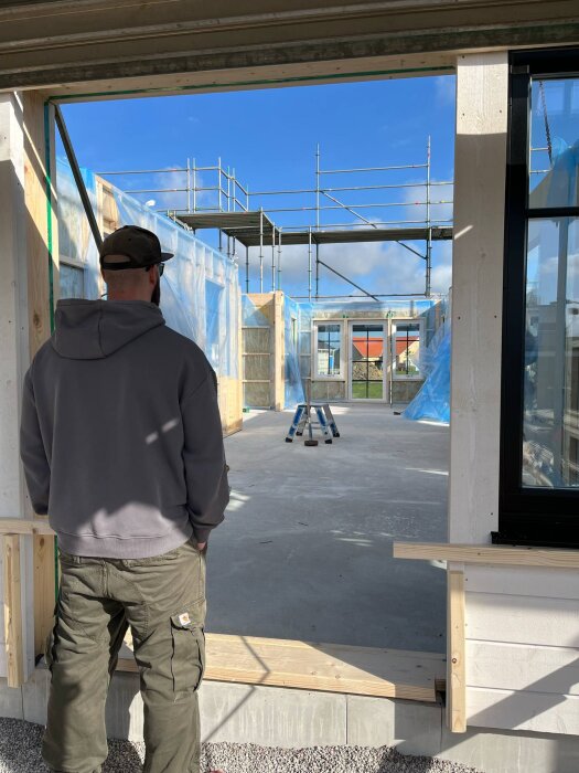 Person inspekterar pågående husbygge, med delvis uppförda väggar och byggnadsställningar, i soligt väder.