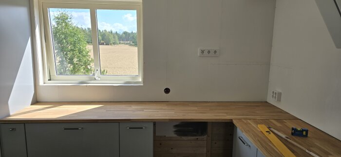 Kök med träbänkskiva, sned vägg och fönster med utsikt över fält och skog. Vattenpass och måttband på bänken.