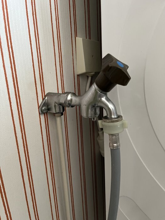 Väggmonterad kran med brun vred mot randig bakgrund, ansluten till en grå slang.