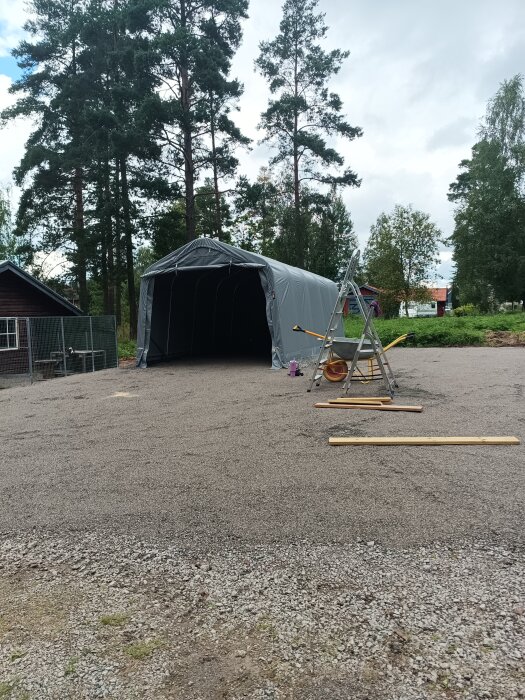 Tältstomme på grusplan under uppbyggnad, omgivet av skog och byggmaterial, stege och träbrädor i förgrunden.