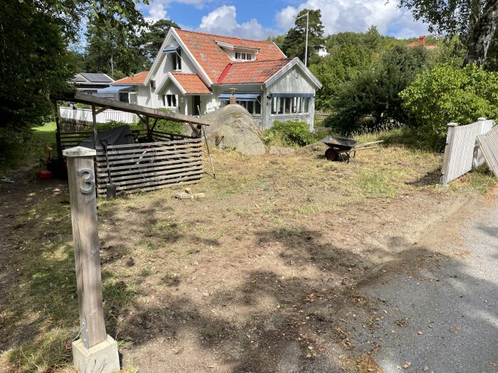 Gammal gräsmatta med stenrik jord, en skottkärra, trästaket, hus med rött tak i bakgrunden, träpelare med siffran 3.