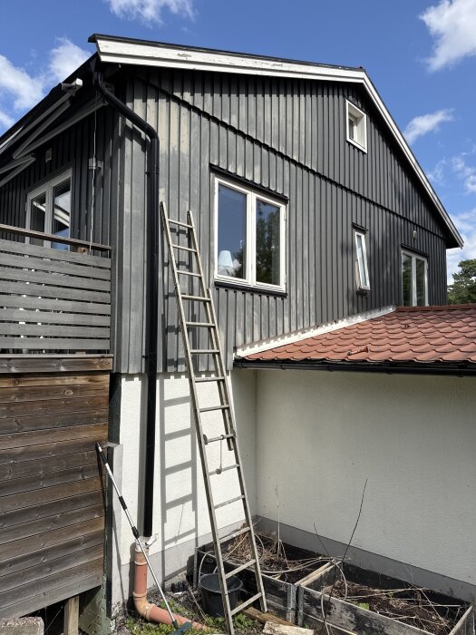 Gråmålad husfasad med fast stege och liten balsäng intill rött tak och träplank. Två fönster syns och växlande molnighet på himlen.
