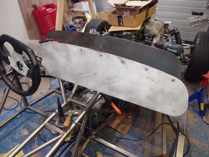 Instrumentpanel påbörjad på bilprojekt, med ratt och skruvhål, omgiven av verktyg och delar i ett garage.