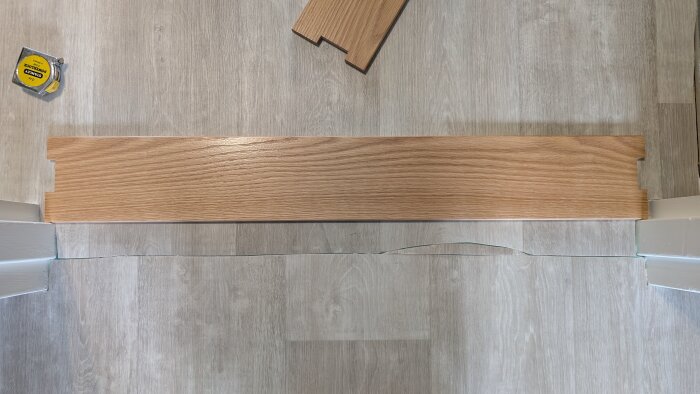 Tröskel av ek ligger på golv i dörröppning, dörrkarm syns. Tröskeln täcker inte hela bredden och ett måttband ligger nära.