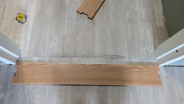 Tröskelbräda i ek som är för kort för dörrkarmens bredd, med ett öppen glipa på ena sidan, liggande på nylagt ljust golv, måttband syns.