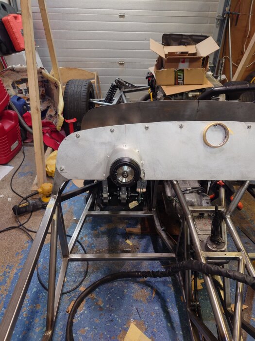 En pågående bilrenovering med metallramverk och kartonglåda på arbetsbänk i garage. Verktyg och bildelar synliga omkring.