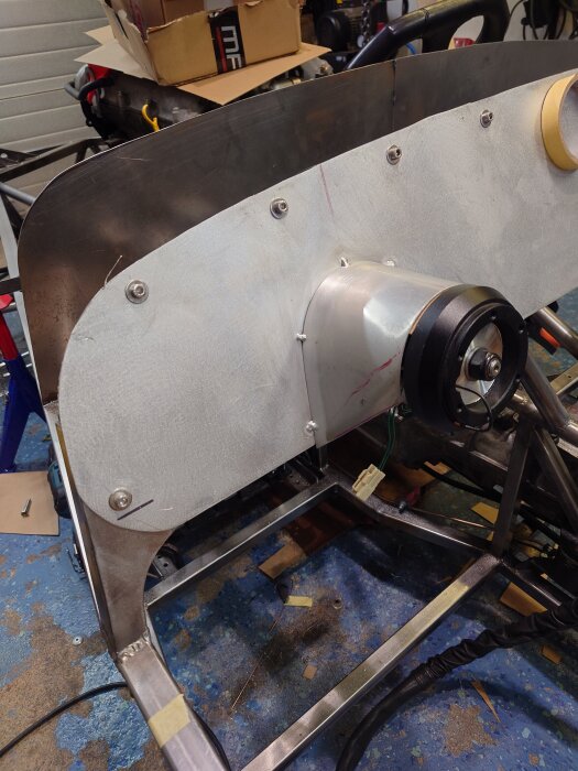 En plåt med bultade fästen och en svetsad ram i ett verkstadsgolv. Kartong och verktyg i bakgrunden.