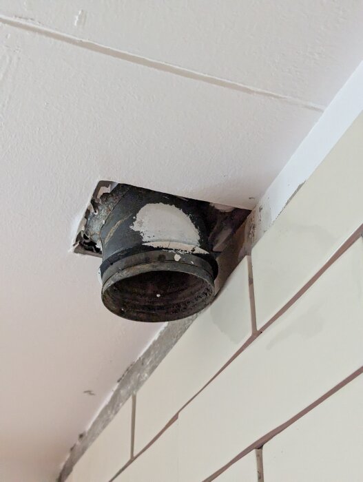 Frånluftsrör på 100mm sitter nära väggen i ett hörn mellan två våningar, omgiven av vita kakelplattor och ett vitmålat tak.