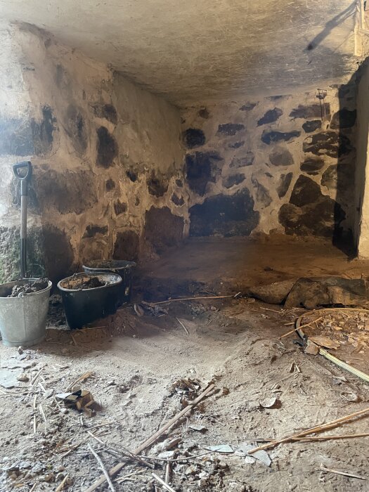 Källarutrymme med stenväggar, jordgolv och byggmaterial. Fyra hinkar står i hörnet vid en spade.