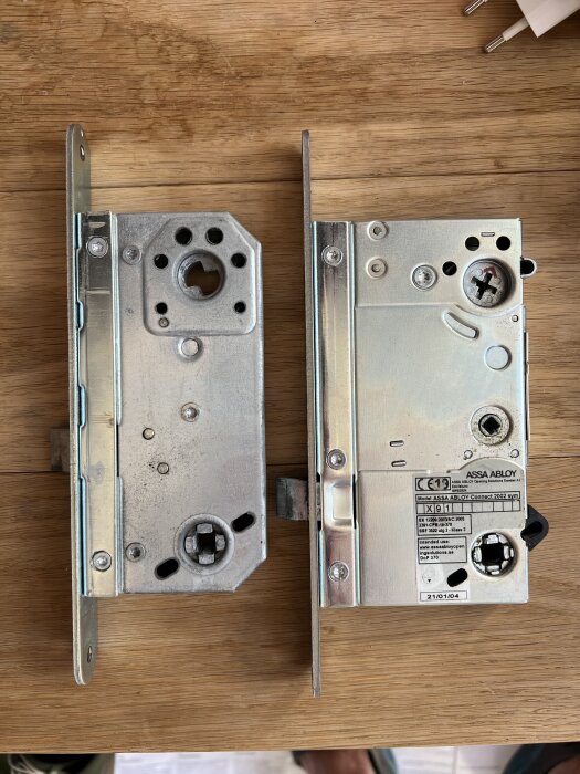 Två låshus av metall, liggande på ett träbord, med olika hål och märkningar, diskussion om storlek.