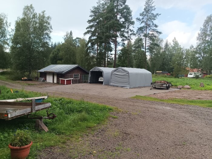 Grusväg med uppställda tält och ett garage i skogsmiljö, omgiven av träd och gräs, samt en släpvagn i förgrunden.