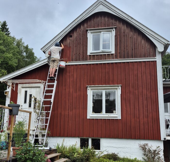 Person målar röd träfasad på ett tvåvåningshus med vit dörr och fönster, med stege och färgburk. Omsorgsfullt arbete pågår.