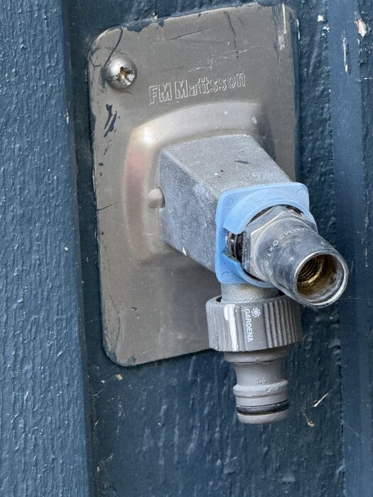 Grå vattenutkastare med en sexkantig detalj och blå tätning, märkt FM Mattsson på en husvägg. Gardena-koppling fäst på kranen.
