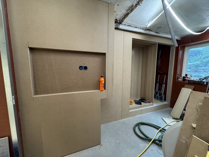 Byggprojekt med MDF-panel och plywood, garderobsdörr och bokhylla under konstruktion, verktyg och limtub i rummet.
