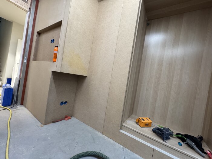 MDF-inklädnad för garderob och bokhylla under byggnation, med verktyg och box vid sidan, på en plywoodyta med pågående installationer.