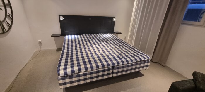 Svart sänggavel gjord av en dörr, med inbyggda ljus och fastsatta sängbord; säng med blåvitt rutigt överkast.