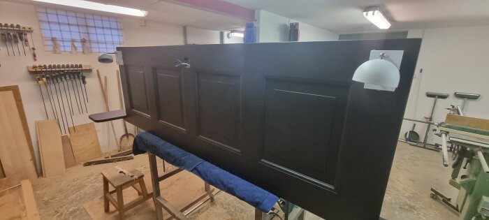 Svart, ombyggd dörr till sänggavel med fast ljusarmatur och sängbord i verkstad.