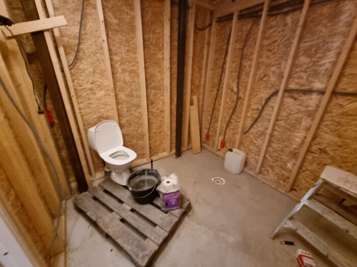 Badrum under konstruktion med synliga träramar, toalettstol, pall och byggmaterial på golvet.