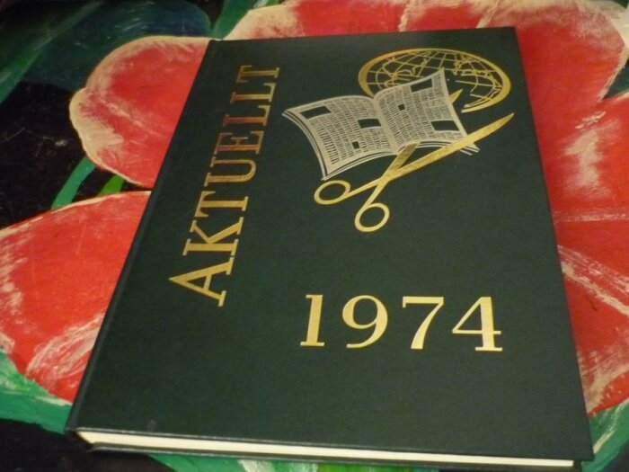 Boken "Aktuellt 1974" med en illustration av en tidning, sax och glob på omslaget, ligger på en färgglad bakgrund.