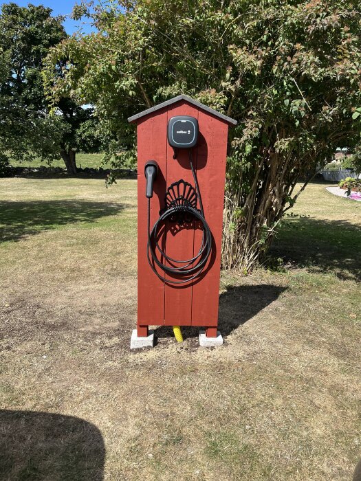 Röd trästruktur i en trädgård med en väggmonterad elbilsladdare, omgiven av gräs och träd.