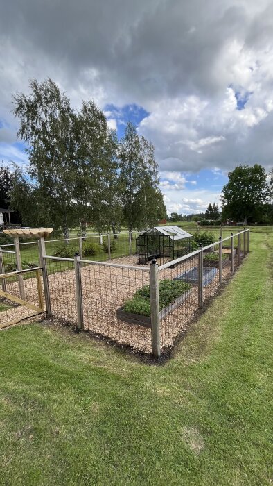 Trädgård med staket, växthus, grusgång och odlingslådor, omgiven av gröna träd och en molnig himmel.