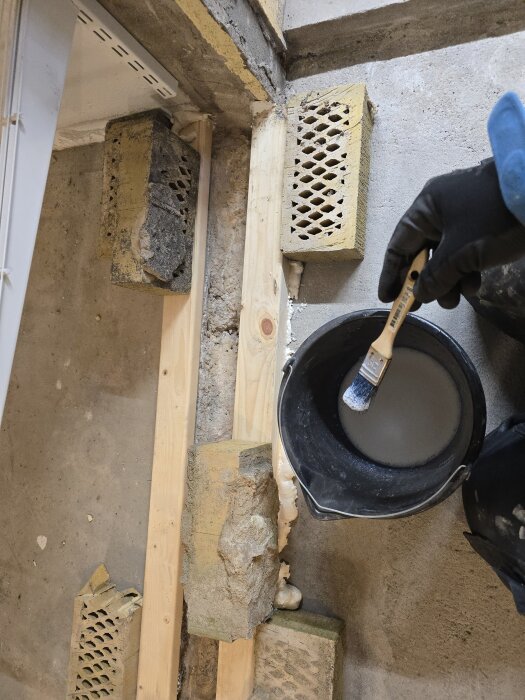 Hand i arbetshandske håller en pensel ovanför en hink. Trä och tegelstenar bildar stöd för en form på ett betonggolv.