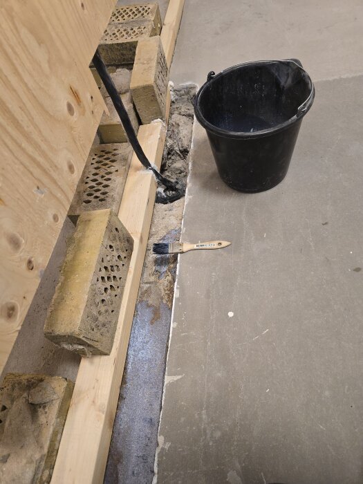 Formvirke och tegelstenar hålls på plats för att gjuta igen en ränna. En pensel och en svart hink syns bredvid på betonggolvet.