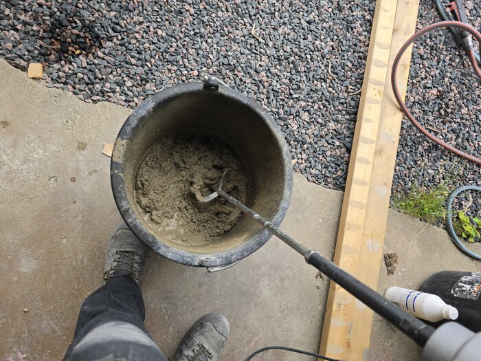 En hink med betongblandning och en omrörare, omgiven av grus och träplankar. En person står bredvid i arbetsskor.