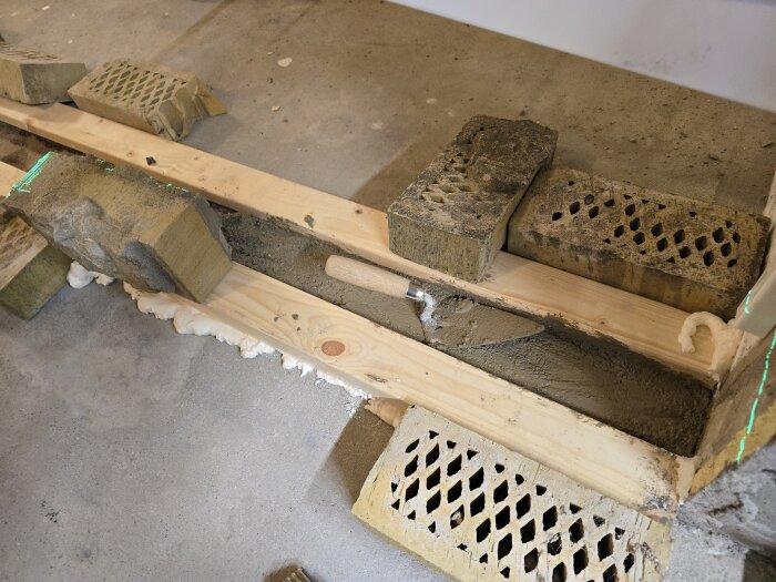 Formbyggnad med tegelstenar och trä för betonggjutning. Betong ligger i formen, fixerad med tegelstenar. Fogskum rundar av kanterna.