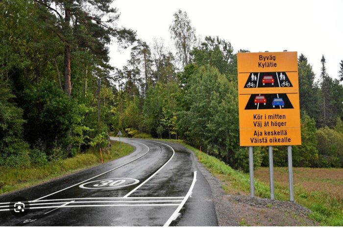 Våt landsväg med en kurva i skogsmiljö, vägskylt visar instruktioner på svenska och finska för trafikanter att köra i mitten och ge plats åt höger.