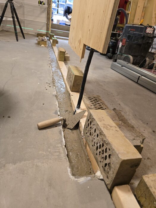 Gjutform för innervägg med betong i en ränna, tegelstenar som stöd och en elkabel genom formvirket. Murarslev ligger vid sidan.