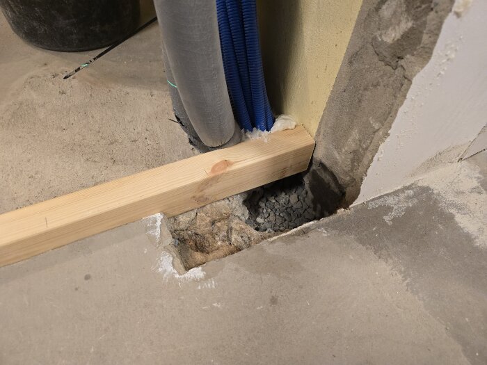 Träplanka stöds med tegel i ränna, blå och grå kablar löper intill. Gjutning av betong pågår för innervägg i renoveringsprojekt.