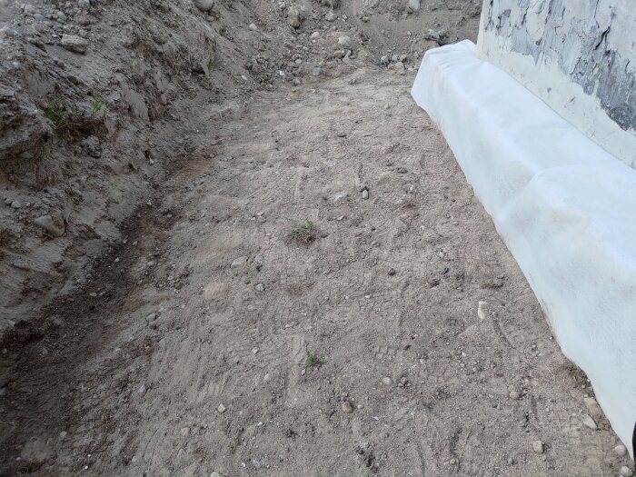 Utgrävd och torr sandig mark vid husgrund, täckt med vit material vid husväggen, synlig pga varmt och soligt väder.