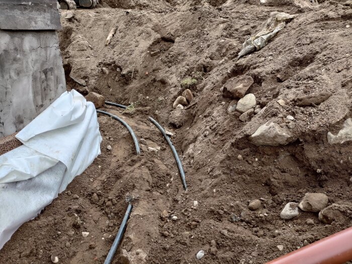 Grävd mark med lagda PEM-slangar och stenar synliga, samt skyddande material vid en betongvägg.