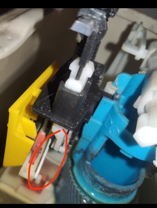 Närbild på en fjäder/vajer markerad med röd cirkel inuti en toalettcistern. Blandning av plastdelar i olika färger.