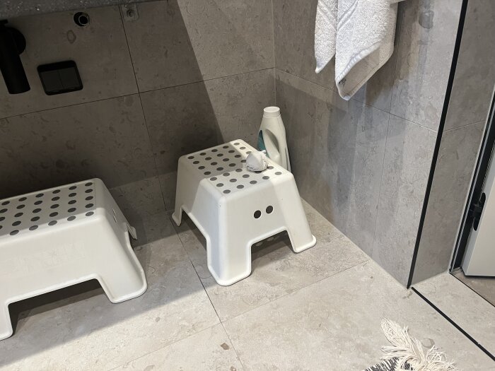 Vit plastpall på badrumsgolv med kakelvägg i bakgrunden, fläckar synliga nära hörnet.