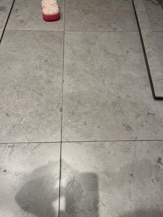 Grå badrumsgolvplattor med vita och rosa rengöringssvampar, synliga foglinjer och mögel eller kalkfläckar.