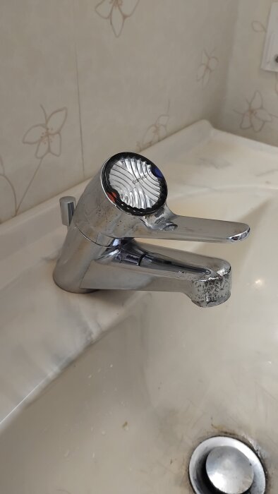 Kromad badrumsblandare från 70/80-talet, märkt Damixa, installerad i en vit handfat med mönstrad tapet i bakgrunden.