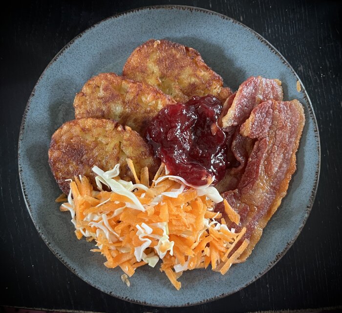 Stekt fläsk, potatisbullar med lingonsylt, serverat med vitkål och morotssallad på en tallrik.